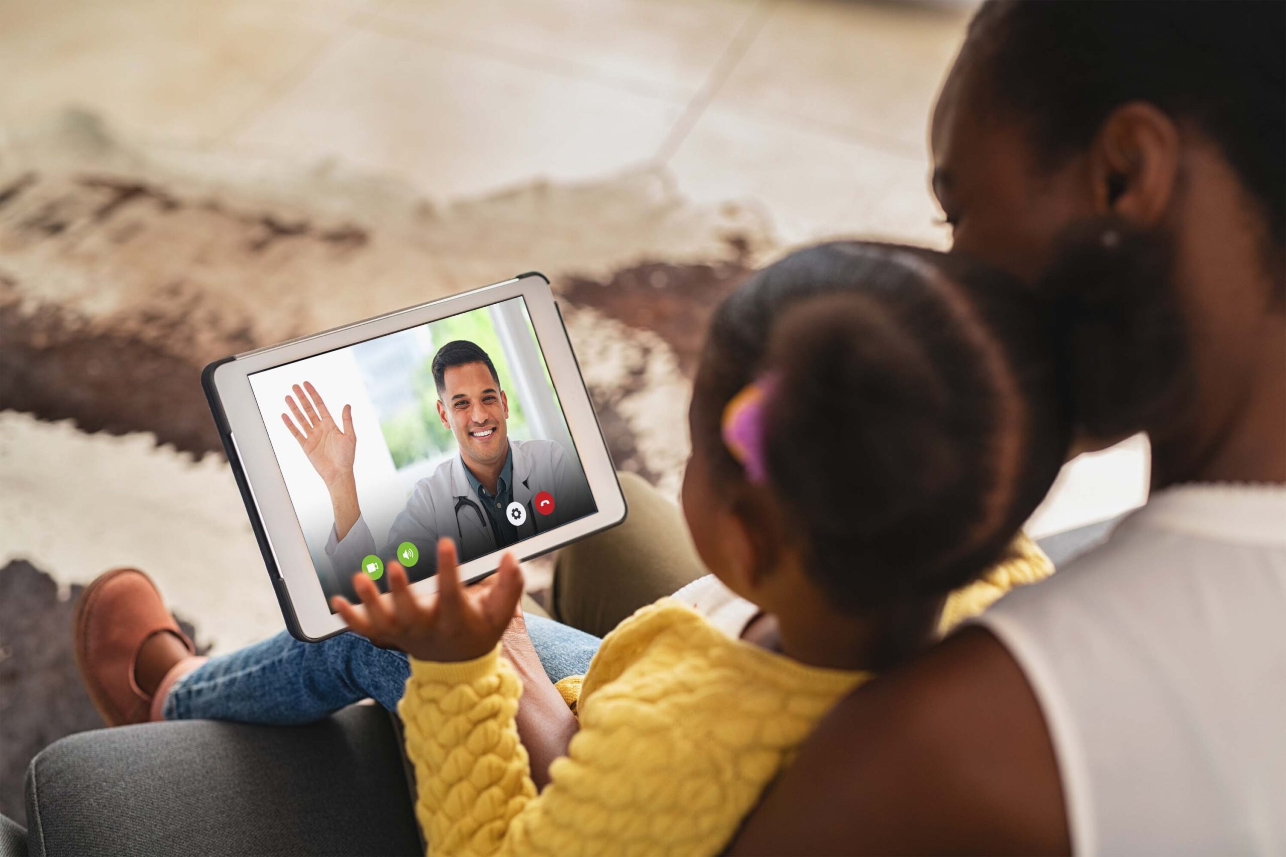 Black mother and daughter on tablet waving to smiling male doctor on a virtual visit.