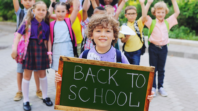 Group of young students with backpacks outside holding chalkboard sign that says "BACK TO SCHOOL".